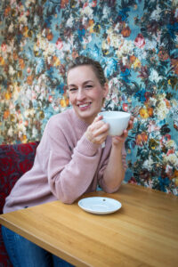 Frau sitzt mit einer Kaffeetasse in der Hand in einem Café und lächelt in die Kamera