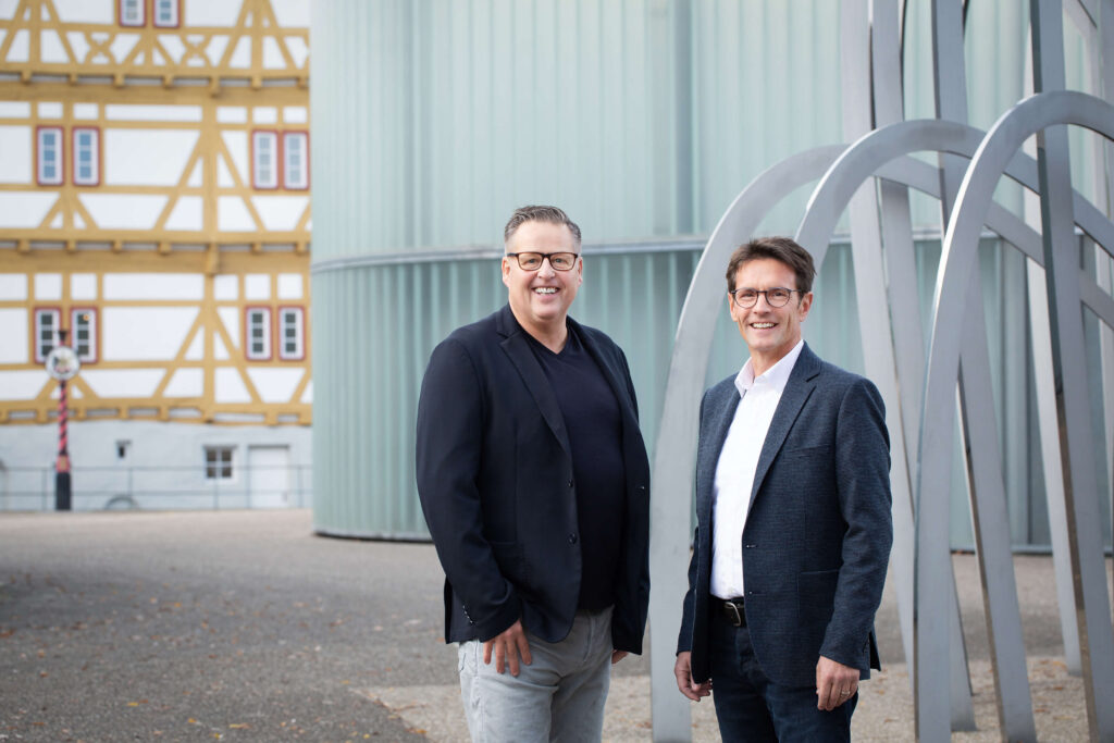 Teampartner-Foto vor modernem Kunstwerk, zwei Männer, im Hintergrund ein Fachwerkhaus, farbig